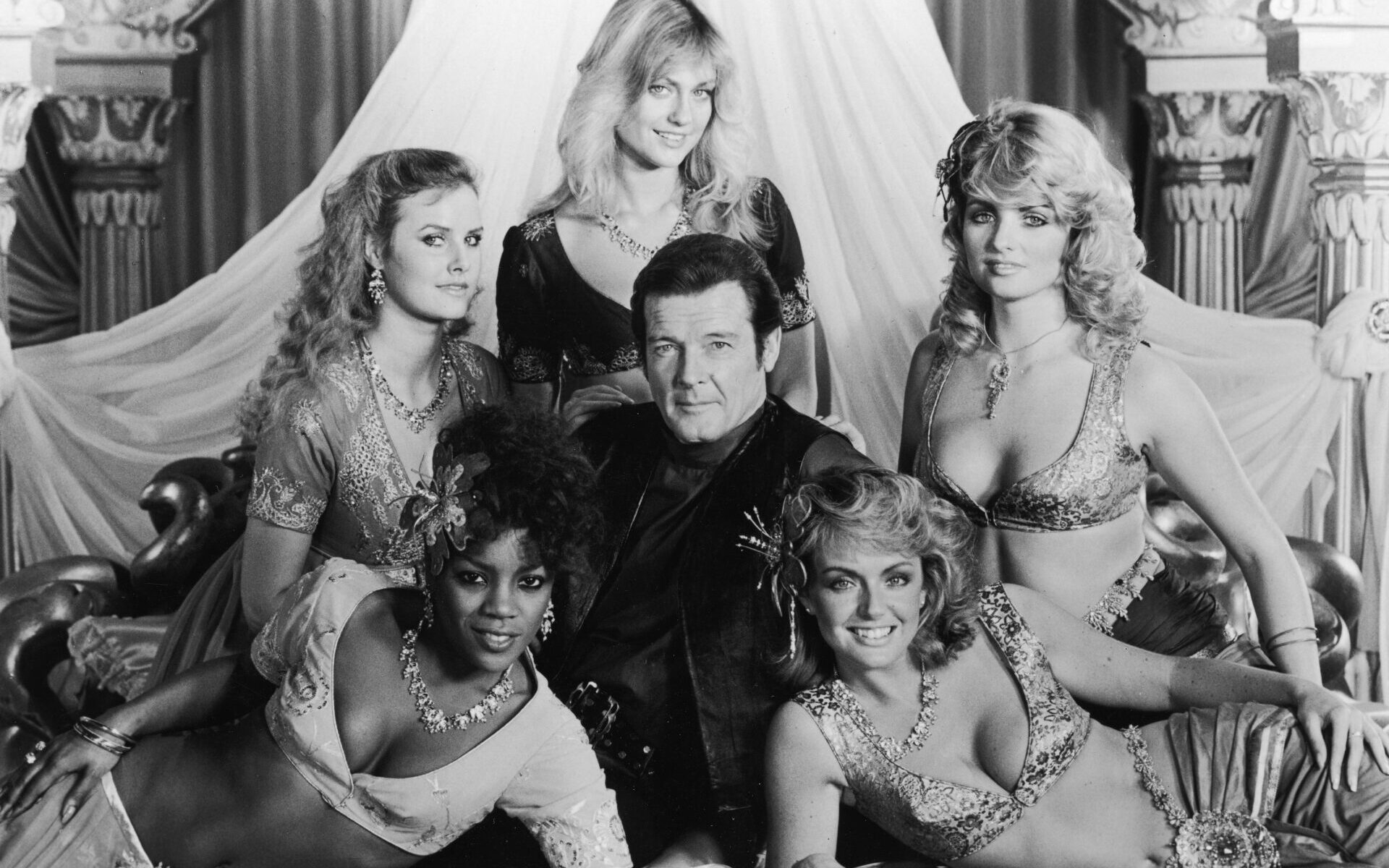 A promotional shot of British actor Roger Moore (as James Bond) surrounded by female palace guards in the film 'Octopussy', 1983. Clockwise from top, they are Carolyn Seaward, Carole Ashby, Tina Robinson, Gillian De Terville and Mary Stavin. (Photo by United Artists/Archive Photos/Getty Images)
